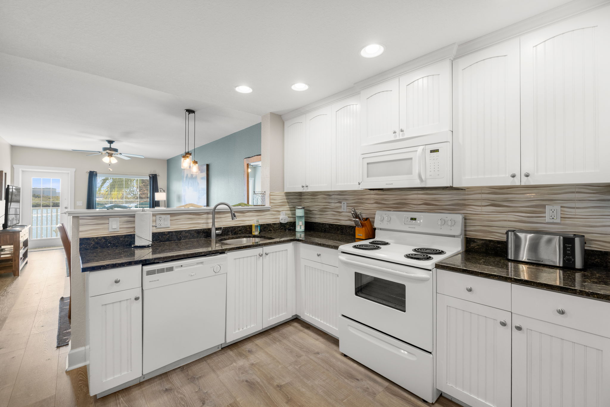 Fully equipped kitchen in Indian Rocks Beach STR condo with white cabinets granite countertops open plan to living area with Intracoastal water view visible through balcony door - well equipped kitchen drives five star guest reviews and longer booking windows