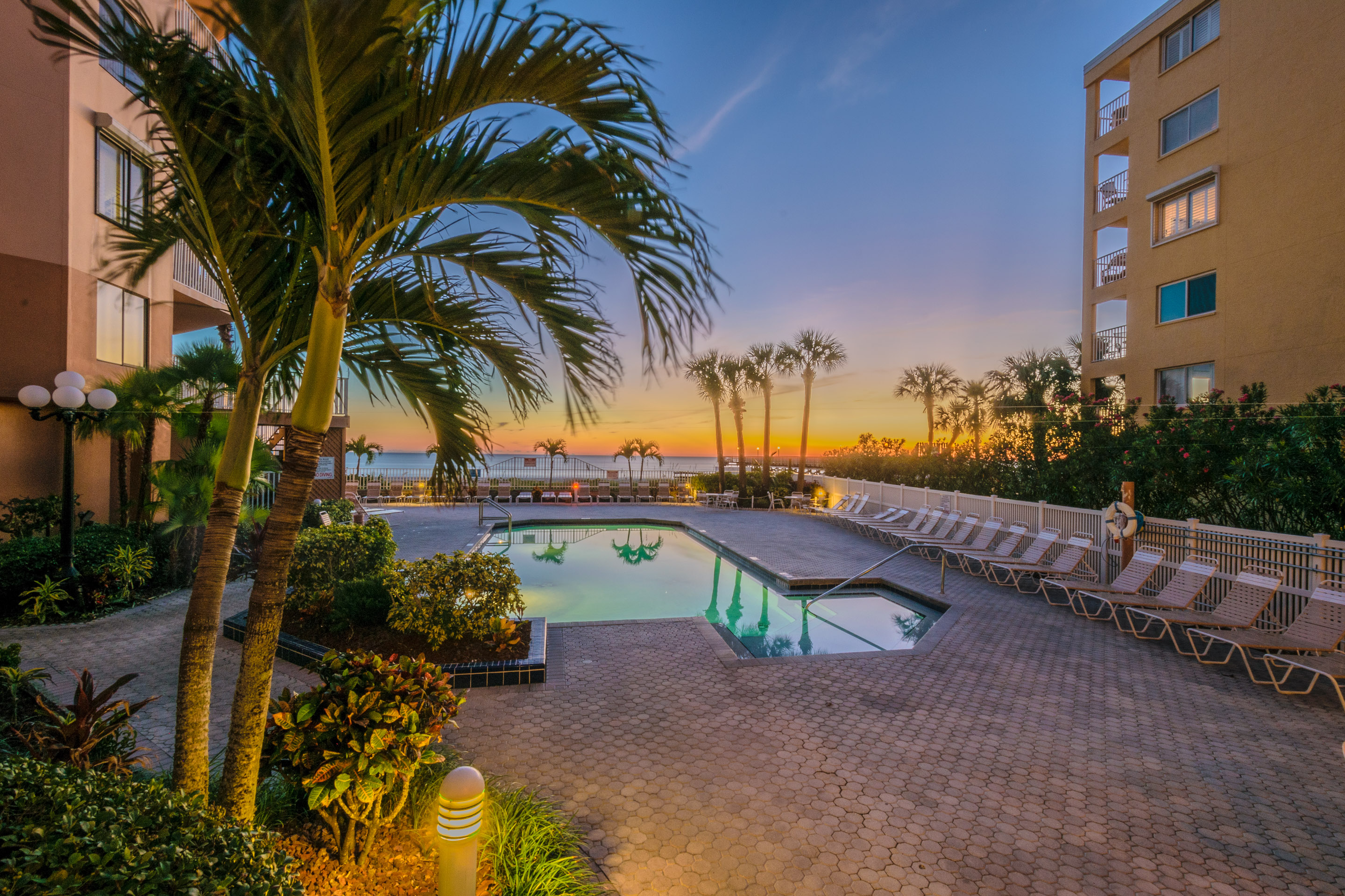Beach Palms condo pool at sunset in Indian Shores Florida Gulf-front vacation rental property with palm trees silhouetted against orange and pink sky Gulf of Mexico visible in background Pinellas County STR investment
