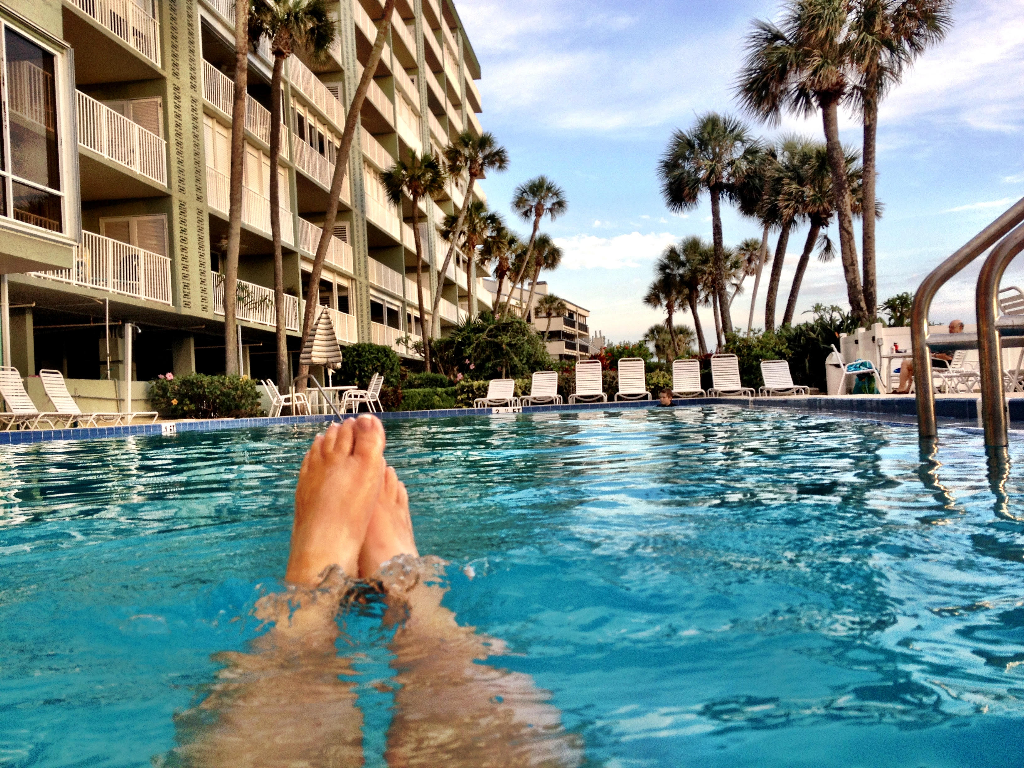 Person relaxing in Gulf-front condominium swimming pool at Sand Castle I Indian Shores Florida with multi-story condo building palm trees and blue sky Pinellas County vacation rental investment property