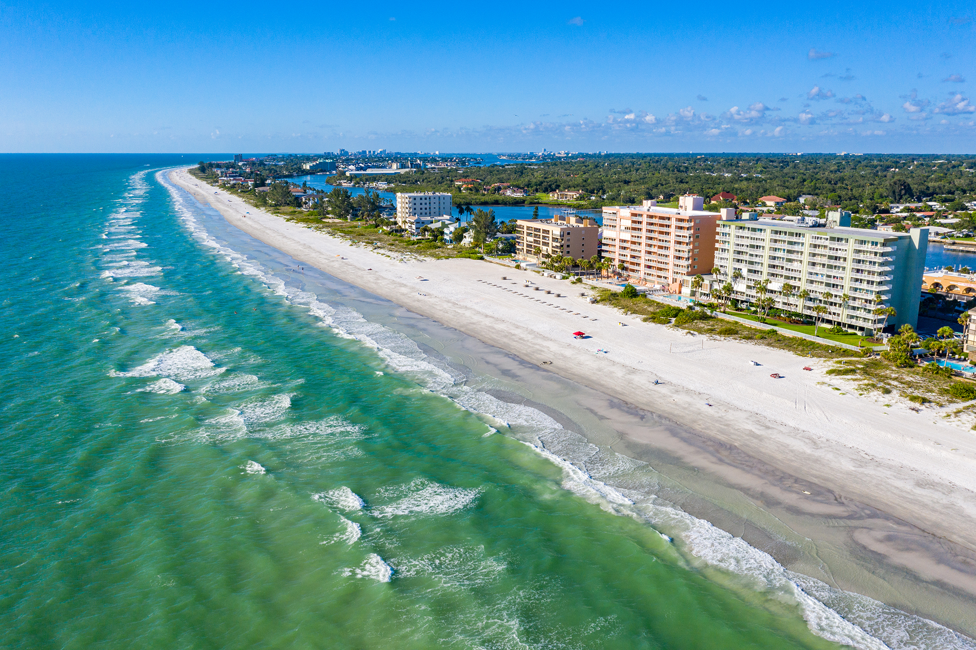 Aerial view looking north along Indian Shores Florida barrier island beach showing Sand Castle I Sand Castle II and Sand Castle III Gulf-front mid-rise condominium buildings along Gulf Boulevard with white sand beach Gulf of Mexico and Intracoastal Waterway visible short-term rental investment properties in Pinellas County