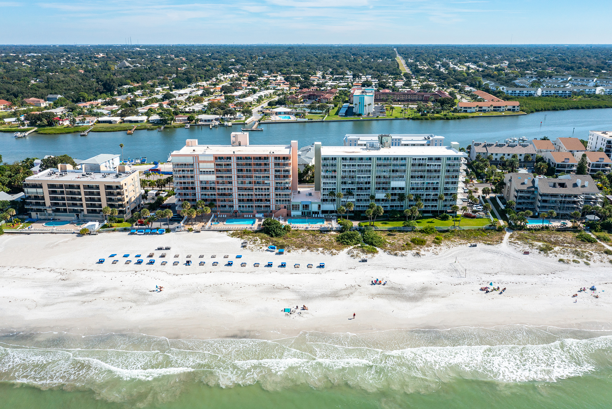 Aerial view of Sand Castle I II and III Gulf-front condo buildings on the beach in Indian Shores Florida with the Gulf of Mexico and white sand beach in the foreground Intracoastal Waterway visible behind STR-eligible vacation rental investment properties Pinellas County