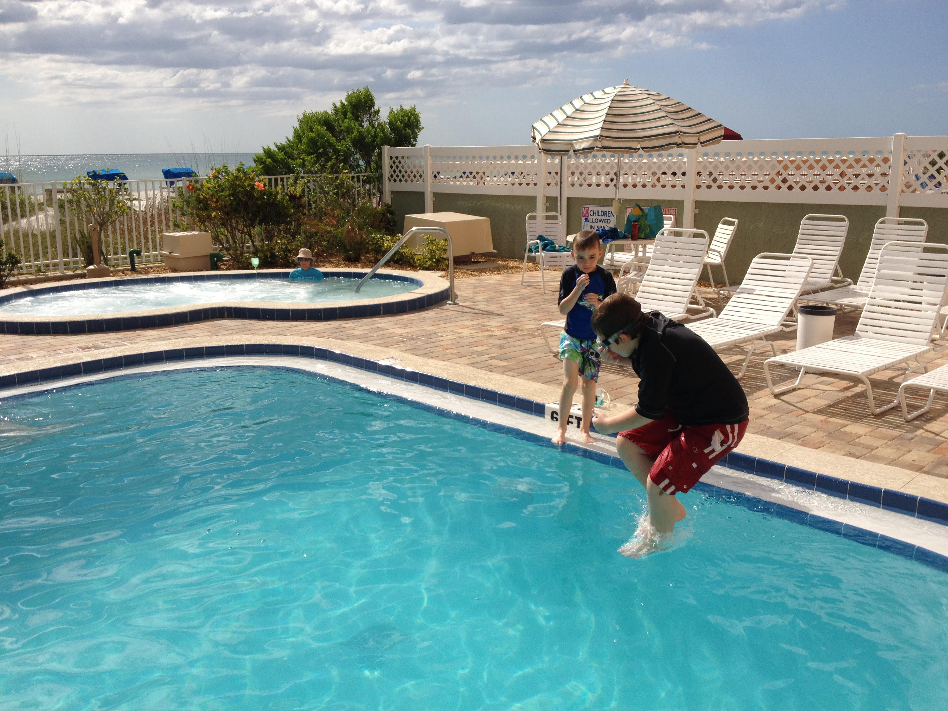 Children jumping into Gulf-front condominium swimming pool at Indian Shores Florida vacation rental property with Gulf of Mexico white sand beach and hot tub visible in background Pinellas County STR investment family memories