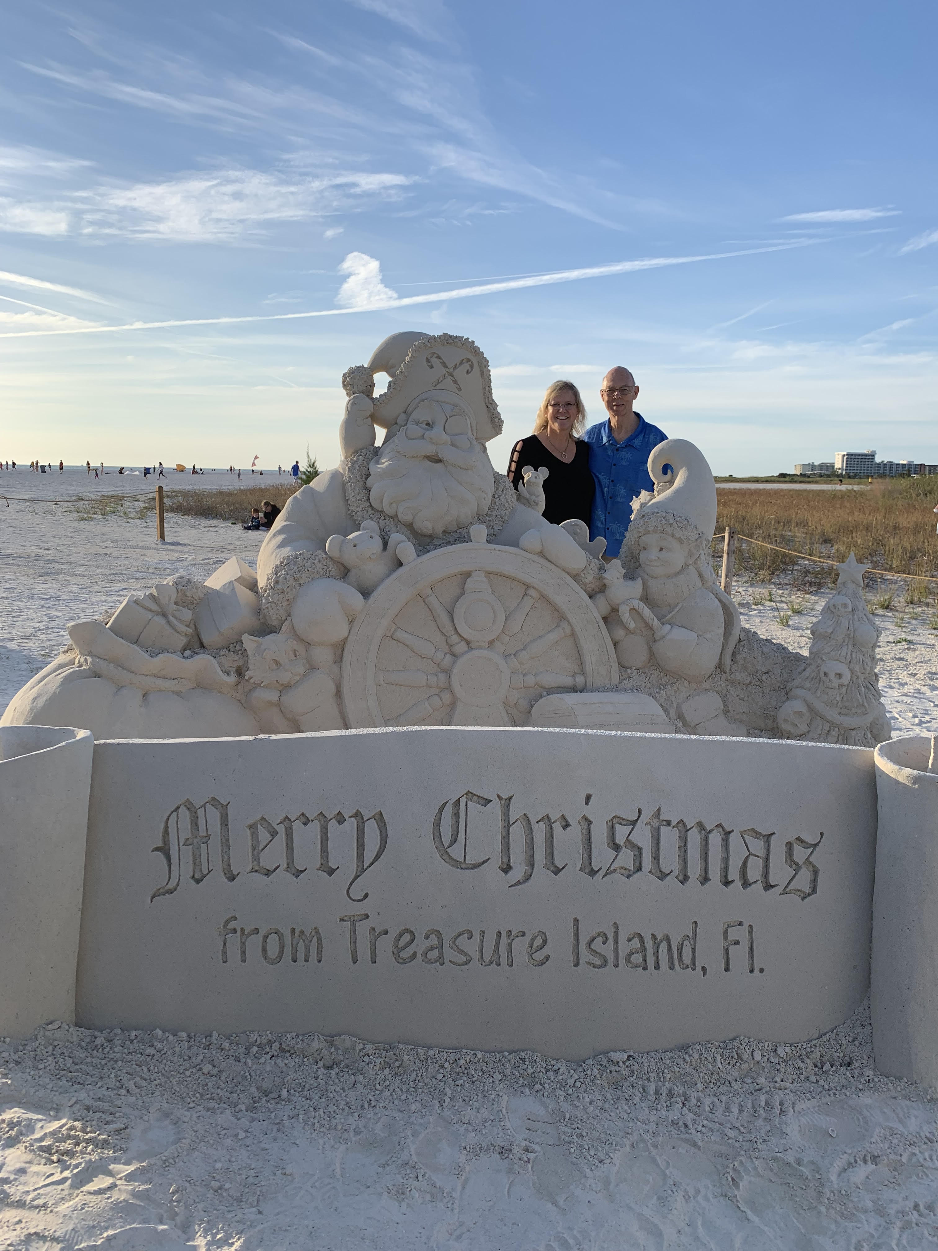 Cyndee Haydon and Jack Haydon Gulf Beaches residents and property owners at Sanding Ovations standing in front of Merry Christmas from Treasure Island Florida sand sculpture Pinellas County
